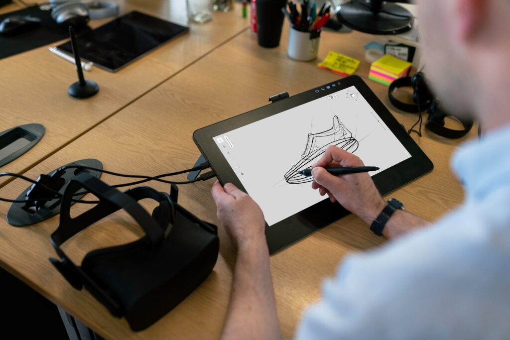Insinööri piirtää tabletilla kengän kuvaa. Työpöydällä, piirtäjän vasemmalla puolella, lepäävät VR-lasit.